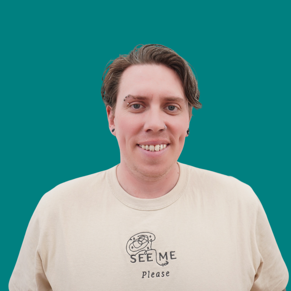 Smiling, with short wavy brown hair and eyebrow and nose piercings, wearing a cream See Me Please t-shirt.