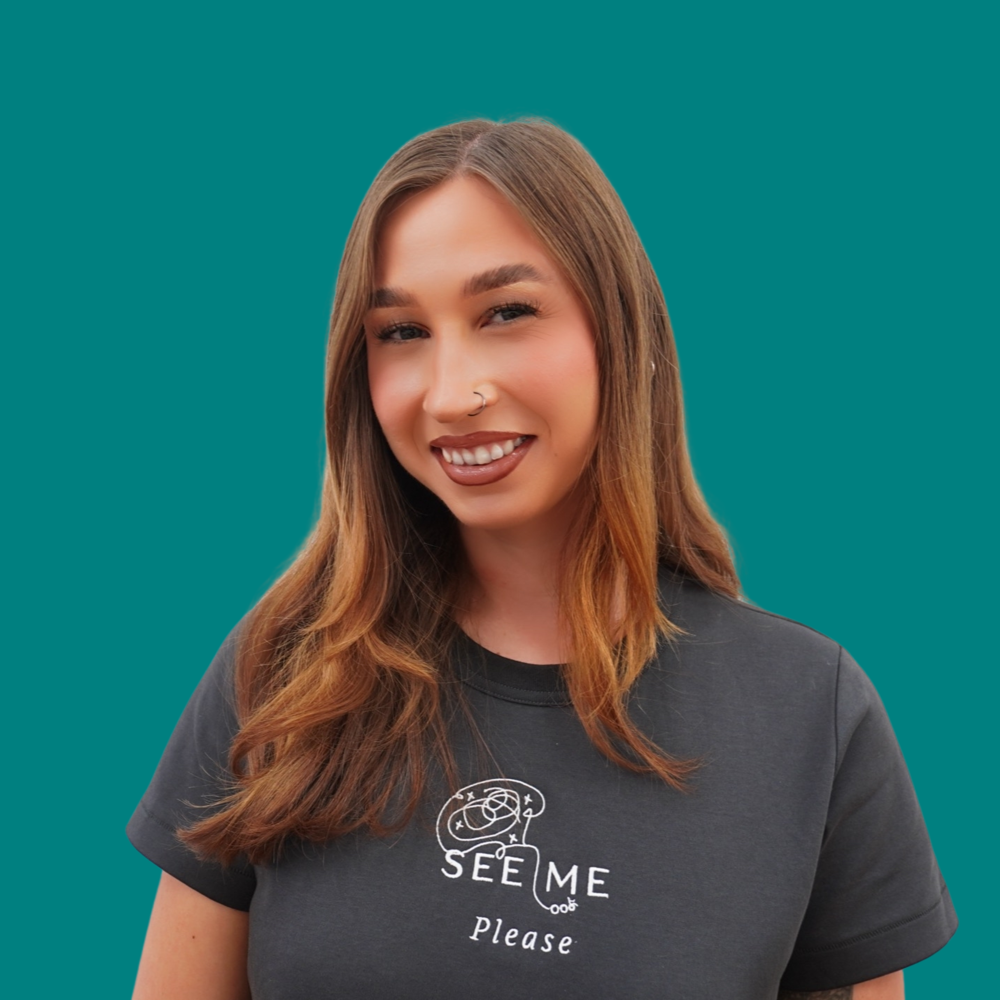 Smiling, with long brown hair and a nose ring, wearing a dark grey See Me Please t-shirt.